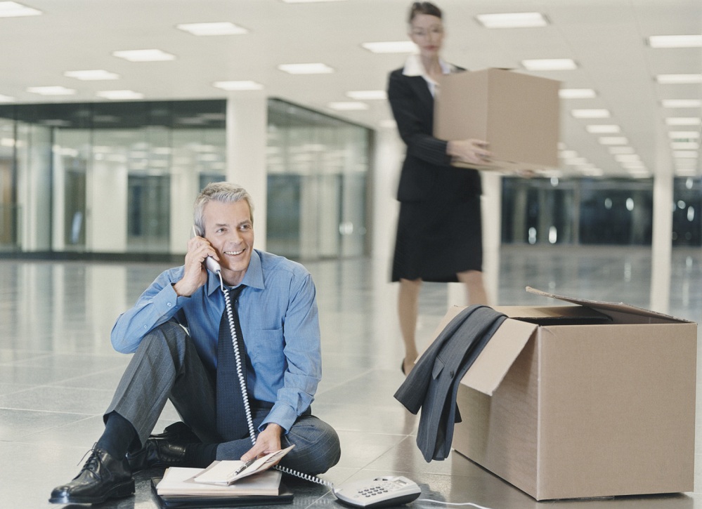 Businessman Sits on the Floor of a New Office, Talking on the Phone, a Woman Carrying a Cardboard Box in the Background